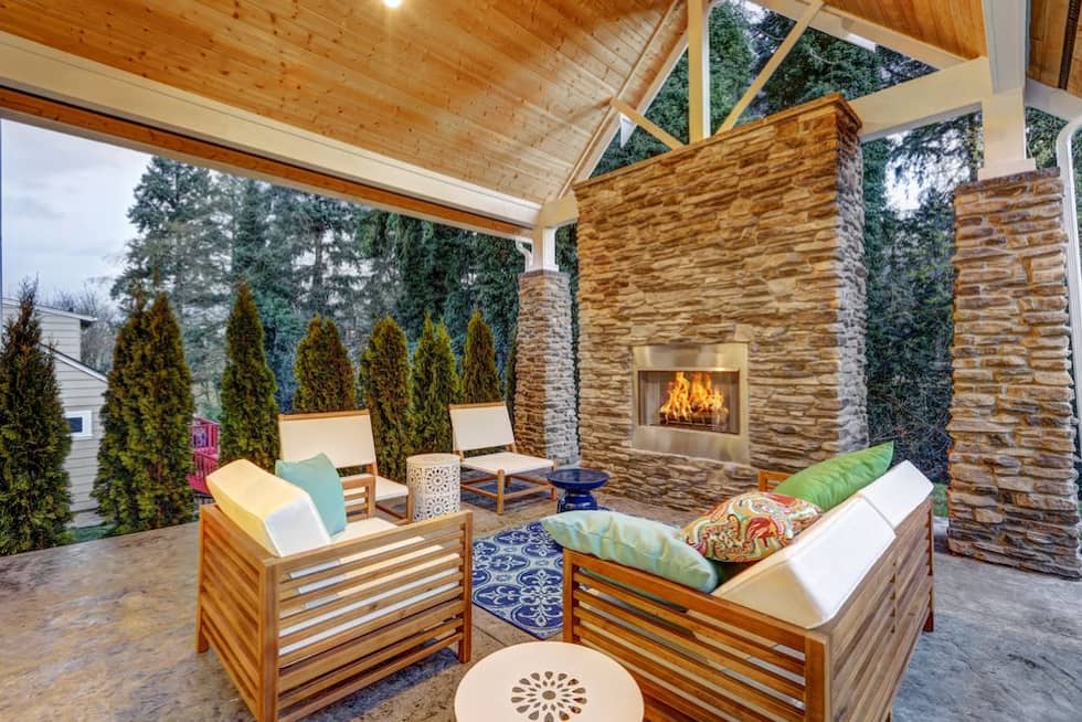 A covered outdoor patio with a built-in gas fireplace, stone cladding, wooden seating, and colourful cushions, surrounded by tall evergreen trees.