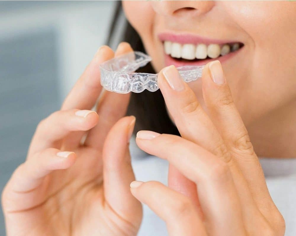 A woman holding a clear aligner near her mouth, highlighting the everyday use and hidden costs of teeth straighteners.