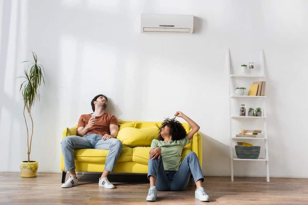A frustrated couple sitting on a yellow couch beneath a heat pump that’s not working properly in a modern home.
